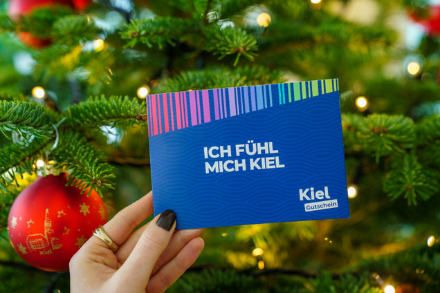 hand hält KielGutschein vorm Weihnachtsbaum